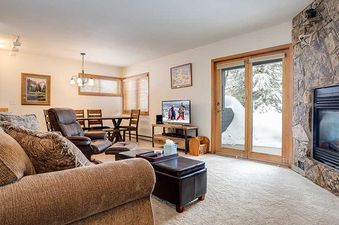 A cozy living room at Pines Condominiums Two Bedroom with a brown sofa, leather recliner, television, and fireplace, with a dining area visible in the background and a snowy outdoor view through a glass door.