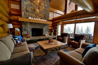 A cozy living room in Totem Chalet featuring a large stone fireplace, comfortable sofas, and armchairs, with a view of snow-covered trees through large windows.