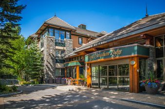 A large lodge with a stone and wood exterior, featuring multiple windows and a prominent entrance with a ‘Royal Canadian Lodge’ sign, surrounded by green trees and flower beds under a clear blue sky.