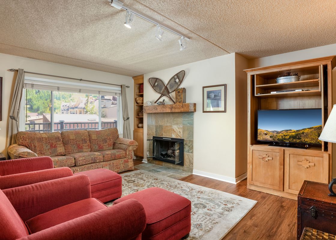 A cozy living room featuring a fireplace, a patterned sofa, red armchairs, a wooden entertainment center with a TV, and a window with an exterior view.