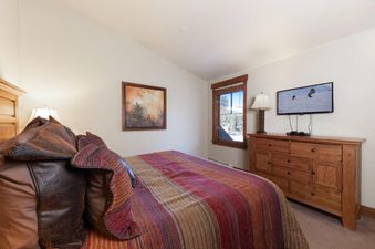 A cozy bedroom with a large bed featuring a colorful striped comforter and dark brown pillows, a wooden dresser with a flat-screen TV showing skiers, and a window with a view of snow-covered trees.