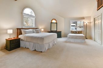 A spacious bedroom at Blue Bird on Norfolk featuring a queen-sized bed, a chaise lounge, nightstands, lamps, and a dresser with a mirror, all set on light-colored carpet under vaulted ceilings.
