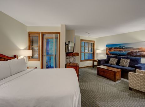 A Blackcomb Springs Premium Studio features a cozy bed, a separate living area with a sofa and armchair, and an in-room fireplace, all brightly lit by natural light from large windows.