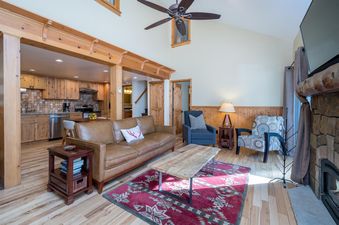 A comfortable and rustic living area with a leather sofa, wooden coffee table, and red area rug, flowing into a modern kitchen with wooden cabinetry and stainless steel appliances.