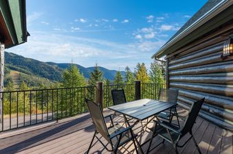 An outdoor deck with a table and chairs, offering panoramic views of forested mountains and a clear blue sky, next to a log-cabin style building.