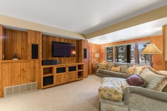 A cozy living room with wood-paneled walls, a large TV, comfortable sofas, and a window overlooking a natural landscape.