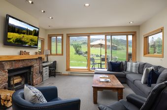 A spacious and inviting living room featuring a large sectional sofa, a coffee table, a fireplace, and a mounted TV, with large windows overlooking a scenic mountain landscape.