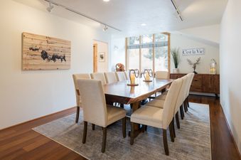 A large wooden dining table sits in the middle of a room with large windows and white walls. The table is surrounded by white chairs, and there are three candle holders with lit candles placed in the center of the table.