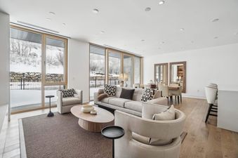 The open-concept living area at Founders Place 2304 features a light gray sofa and armchairs, an oval coffee table, and large windows with snow-covered views.