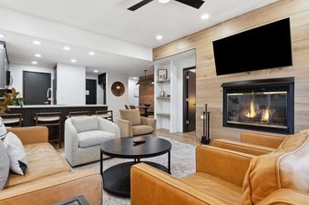 A modern and cozy open-concept living room with leather sofas, armchairs, a black coffee table, and a fireplace with a TV mounted above, next to a contemporary kitchen.