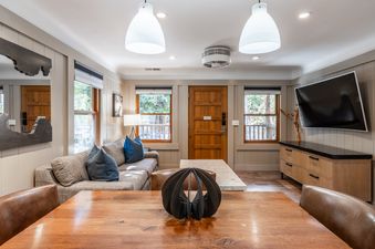 The open concept living area features a comfortable sofa, a large flat-screen TV, a dining table, and large windows with outdoor views.