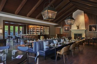 An expansive dining area within the Montage Two Bedroom Alpine Suite, featuring a sophisticated bar area with a rich selection of spirits, a grand stone fireplace, and elegantly set tables, all under a vaulted wooden ceiling.
