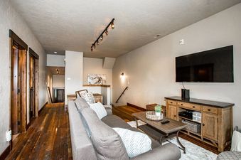 A cozy living room at Cana Loft 302 features a gray sofa, a rustic wooden coffee table, a flat-screen TV mounted above a console, and dark hardwood floors.