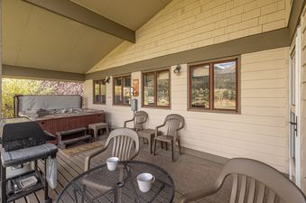 A covered outdoor patio at Glenn House featuring a hot tub, BBQ grill, and an outdoor dining set with scenic views through the windows.