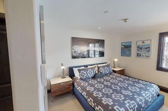 The bedroom at Manor Vail 284 features a king bed with a patterned comforter, two nightstands, a large window with a view of the trees outside, and artwork on the walls.