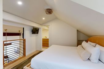 A cozy bedroom loft in Hollyhock 2023 with a white bed, sloped ceiling, woven ceiling fan, and a view into an adjacent area with a wooden railing and a television.