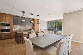 An open concept kitchen and dining room in Founders Place 2303 features modern appliances, a large dining table with upholstered chairs, and a view of a forest through expansive windows.