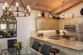 Top of the Village Slope 408 features a well-equipped kitchen with a granite countertop and stainless steel appliances. The kitchen is open to the dining area, with a glass table and four chairs. The unit has a vaulted ceiling and a rustic feel.