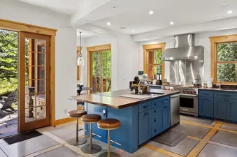 A spacious kitchen with a large blue island, a live-edge wooden countertop, three bar stools, and stainless steel appliances.
