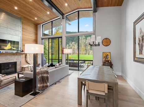 A spacious, open-plan living and dining area with high ceilings, large windows overlooking a golf course and mountains, a fireplace with a TV above it, and modern furnishings.