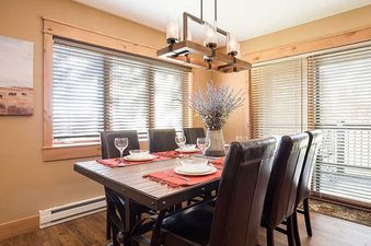 A rustic dining room at Ranch at Steamboat RA301, featuring a wooden table set for six, illuminated by natural light from large windows with blinds and decorated with a vase of purple flowers and a landscape painting.