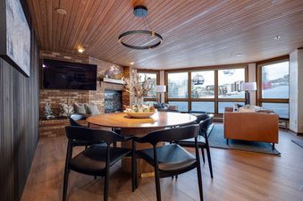 A spacious open-concept living area with a round dining table, a cozy lounge area, a brick fireplace, and large windows offering views of a snowy landscape and ski gondolas.