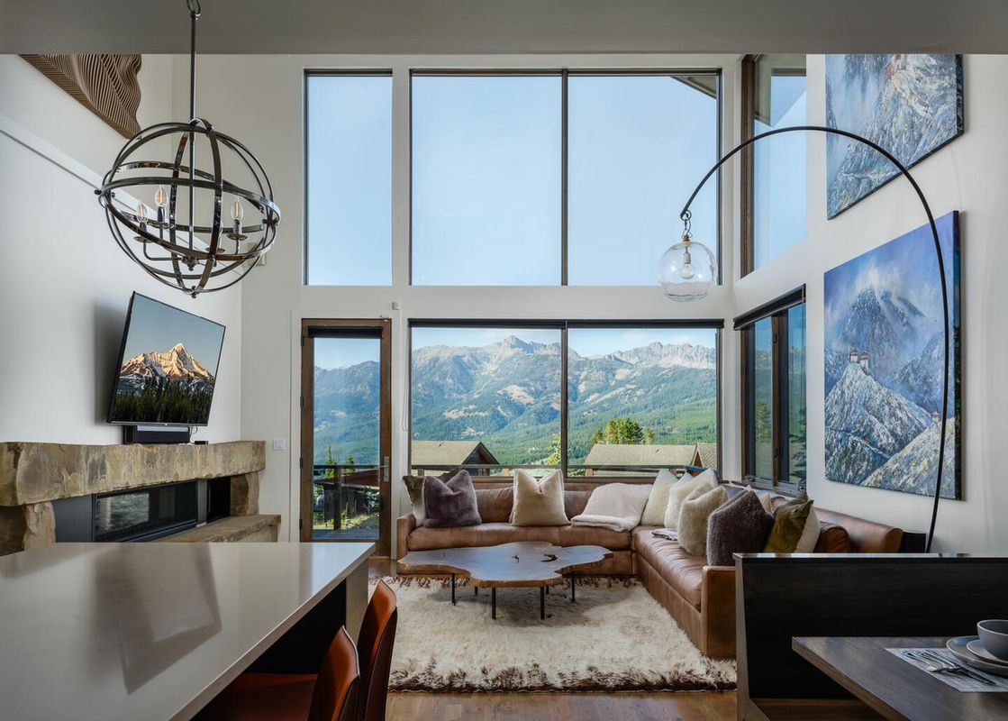 The living room of Moonlight Basin 6 Silver Peaks features a comfortable leather sectional sofa, a large window with mountain views, a fireplace, a large flat screen TV, and a large dining table.