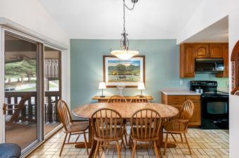 A dining area with a wooden oval table and six chairs, a console table with lamps and a painting, and a partial view of a kitchen and a glass sliding door leading to a balcony.