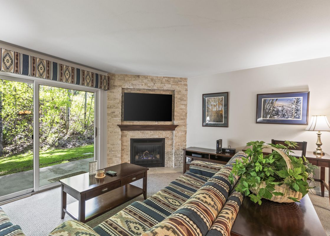 A cozy living room at Snow Flower 135 Park City featuring a comfortable sofa, a dark wooden coffee table, a stone fireplace with a mounted television, and large sliding glass doors opening to a green outdoor view.