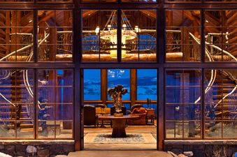 A large, rustic log cabin-style home with expansive windows showcasing a warm, inviting interior and a twilight exterior view of snow-covered mountains.