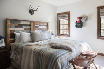 A rustic bedroom featuring a bed with a reclaimed wood headboard, striped bedding, and faux fur throw, with two windows offering natural light.