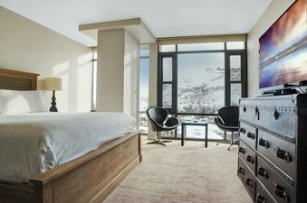 A spacious bedroom at Lowell 301A with a large bed, trunk-style dresser, flat-screen TV, and floor-to-ceiling windows overlooking snowy mountains.