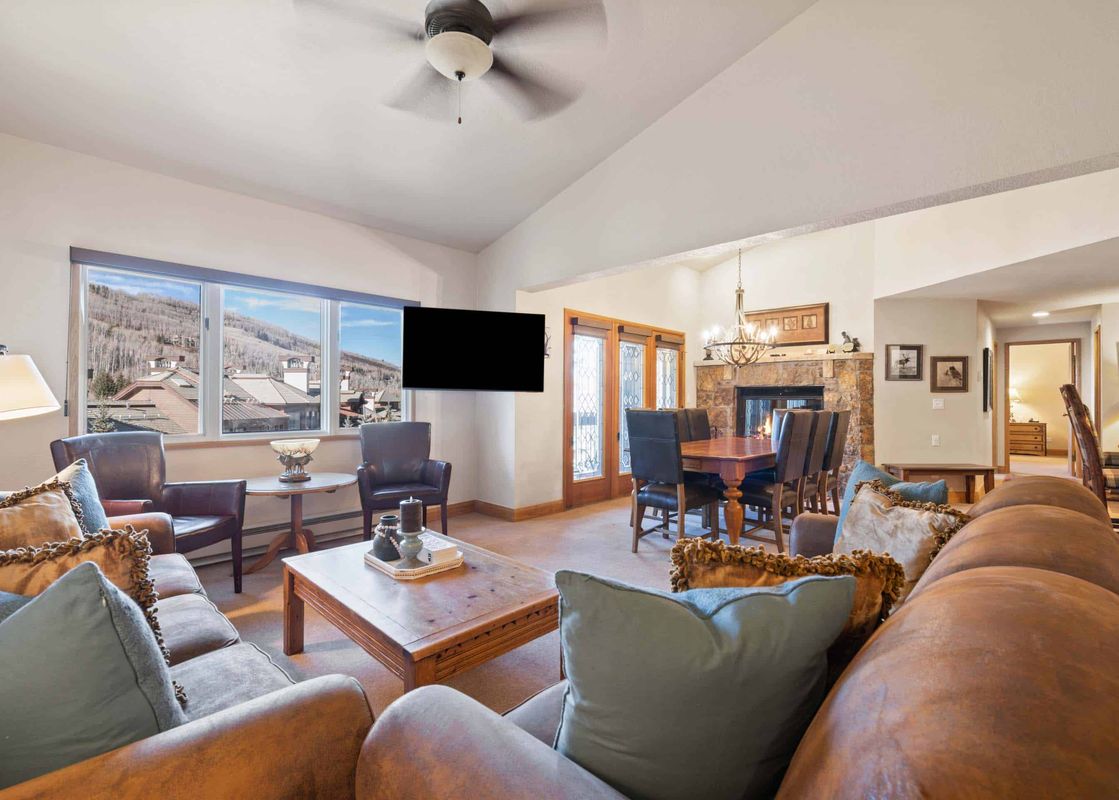 A cozy living room at The Charter F4405 features leather sofas, a wooden coffee table, a flat-screen TV, and large windows offering mountain views, with a dining area and stone fireplace nearby.