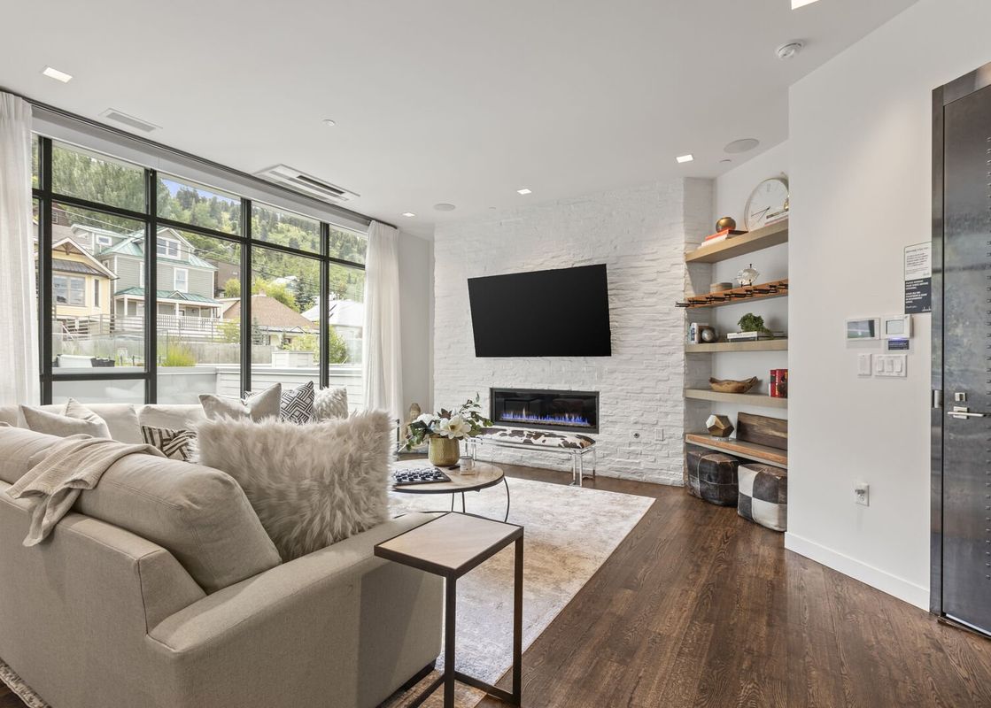 A modern living room at Parkite 37 with a comfortable sofa, a large TV mounted on a white brick wall above a fireplace, and floor-to-ceiling windows offering scenic views of a mountainous neighborhood.