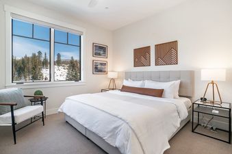 The bedroom is spacious with a large window overlooking a snowy landscape with pine trees. The room features a comfortable bed, a lounge chair, and tasteful decor.