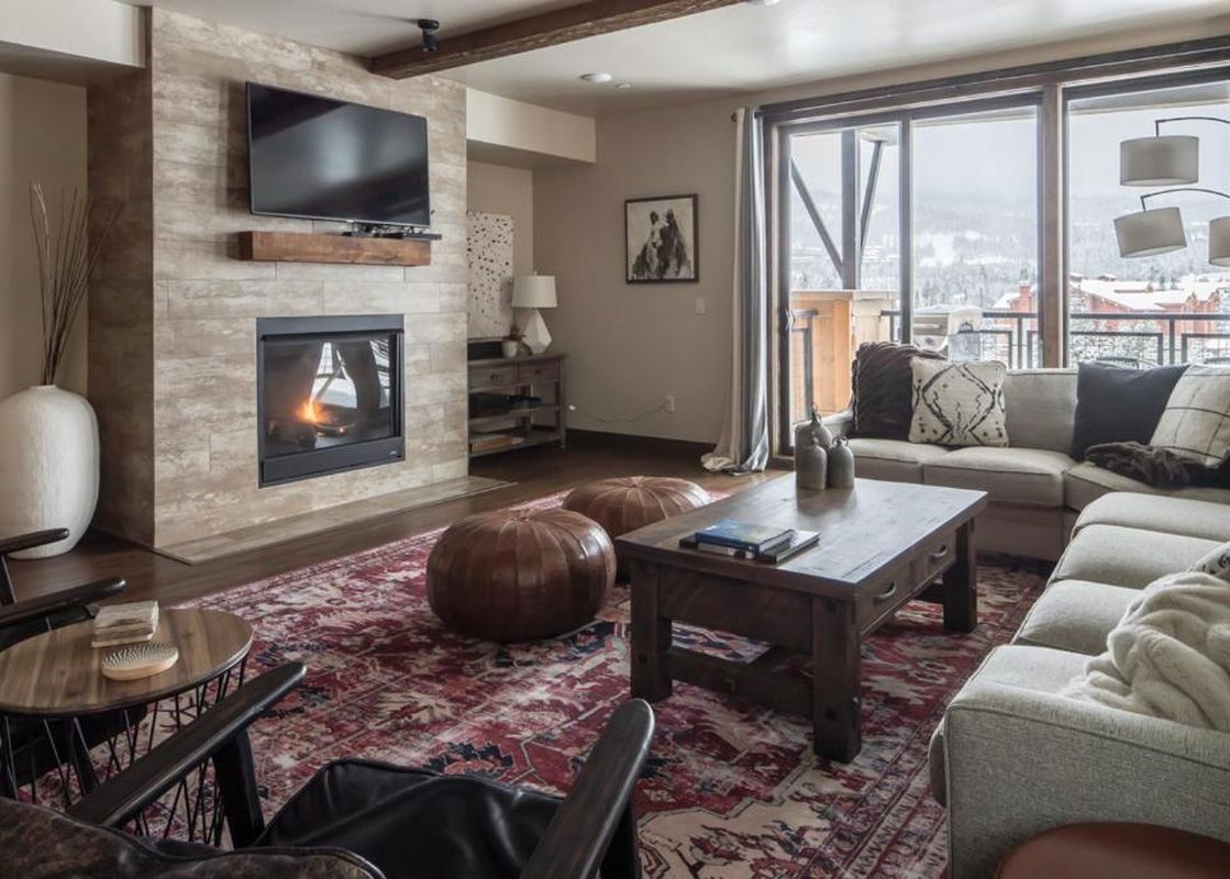 A cozy living room at Mountain Lake Condo 401B featuring a fireplace with a mounted TV, a large L-shaped sofa with decorative pillows, a rustic coffee table, and two brown leather poufs, all set on a patterned rug with a snowy mountain view from the windo