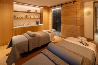 A luxurious spa room in The Shooting Cabin with two massage beds, a vanity with products, and a modern shower with pebble-stone tiling, creating a tranquil retreat.