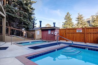 An outdoor pool and hot tub area at Snowcrest 300, surrounded by evergreen trees and a wooden fence, with clear blue water under a bright sky.