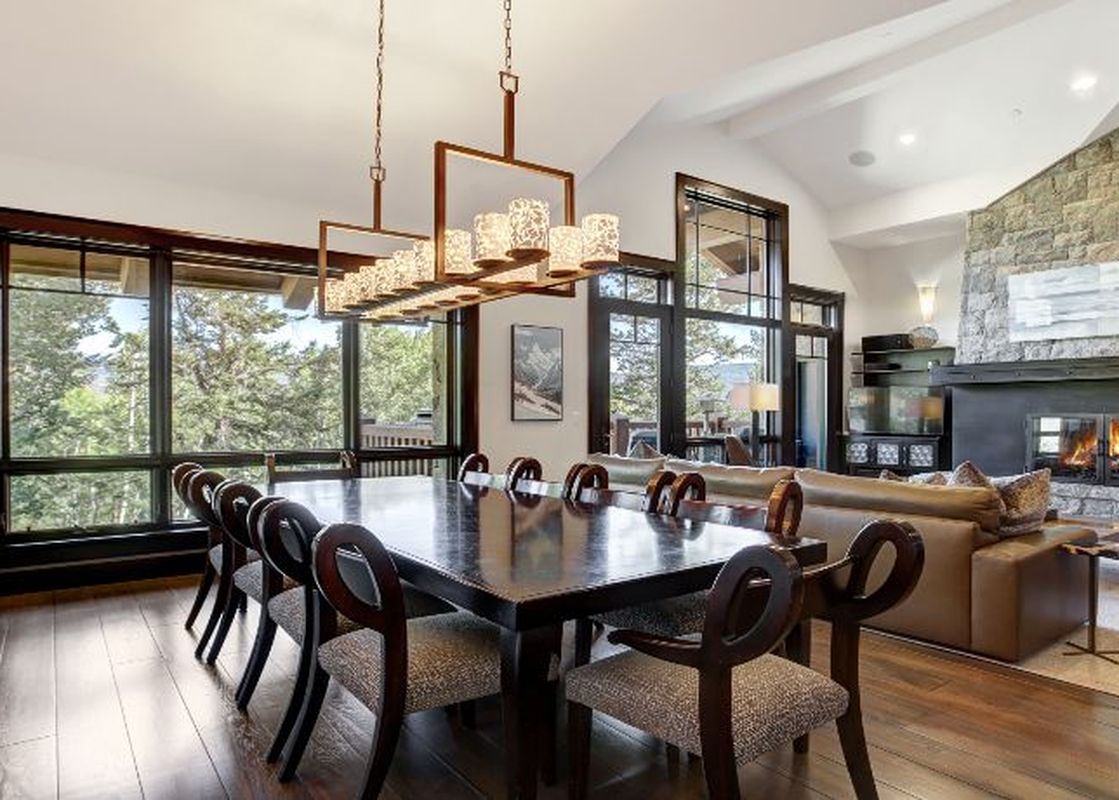 A wide-angle shot of the dining and living area in the Hummingbird Platinum 3 BR Plus Loft, featuring a large dining table with elegant chairs, a modern chandelier, a stone fireplace, and large windows with an outdoor view.