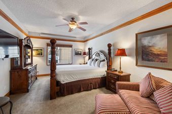 The master bedroom at The Charter F2105 features a four-poster bed, two nightstands with lamps, a dresser with a television, and framed artwork.