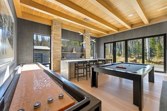 A luxury game room at Kestrel at the Colony, featuring a pool table and shuffleboard, a modern bar, and large windows with scenic outdoor views.