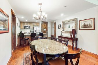 A spacious dining room and kitchen with a granite-topped oval dining table, dark wood chairs, a breakfast bar, and stainless steel appliances.