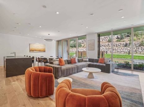 A modern living room with a large grey sectional, orange armchairs, a white coffee table, and expansive windows offering a view of outdoor rock walls and greenery.