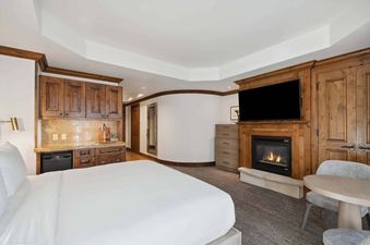 A cozy bedroom in the Aspen 44A features a burning fireplace beneath a wall-mounted TV, a wet bar with wooden cabinets, and a comfortable bed with white linens.
