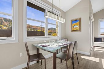 A modern dining area with a glass-top table set for four, featuring brown leather chairs, under a rectangular chandelier, with large windows overlooking autumn mountain scenery.