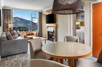 A luxurious living area in The Westin Resort One Bedroom Mountain View features a cozy fireplace, comfortable seating, a dining table, and a large window offering stunning mountain views.