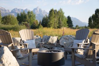 An inviting outdoor fire pit surrounded by four Adirondack chairs, each with a pillow, set against a backdrop of mountains and fields under a blue sky.