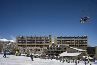 A wide shot of a ski resort on a bright sunny day, showing a large hotel building, a ski slope with many skiers, and a chairlift with people riding it.