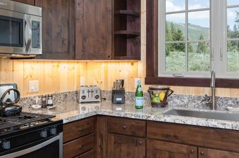 A well-appointed kitchen in The Shooting Cabin features dark wood cabinetry, granite countertops, stainless steel appliances, and a window overlooking a mountain landscape.