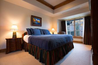 A luxurious king bedroom at Chateau Beaver Creek Residence 406 featuring a king-sized bed with a blue duvet and striped bed skirt, flanked by wooden nightstands with lamps, a window with colorful curtains overlooking autumn trees, and a tray ceiling with